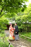 新緑がきれいな庭園で和装の婚礼前撮り写真を撮る新郎と生花ブーケを持つ赤い色打掛の新婦